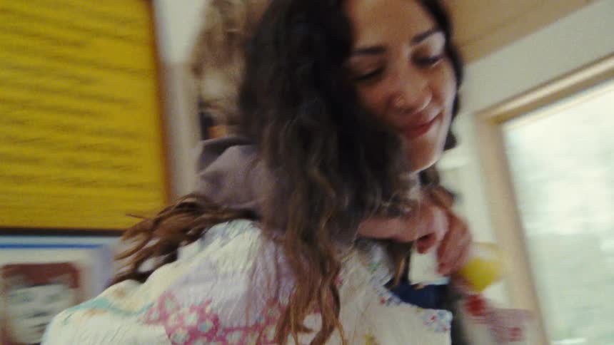 A woman with long dark hair is seen holding a child with short light colored hair on her back as they both smile and she dances and jumps through a brightly lit living room