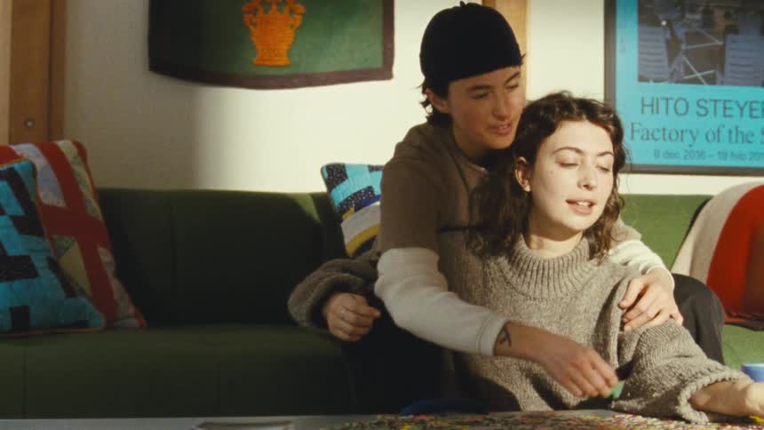 A couple is seen sitting on a couch together the man has his arm around the womans shoulder as they look down and work on a puzzle