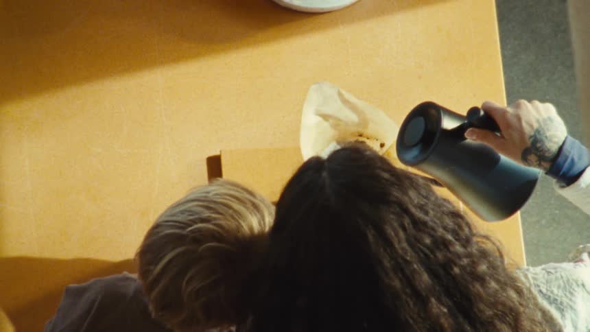 A topdown view shows a person making a pourover coffee the hand with a tattoo holds a kettle to pour hot water over a coffee filter which is placed inside a ceramic mug