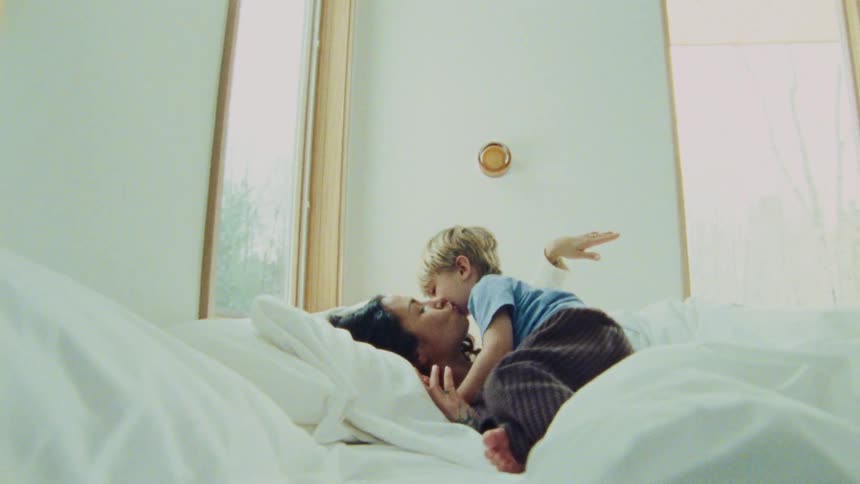 A mother with tattoos on her arm lies in bed and talks with a child with light colored hair then smiles and kisses the child