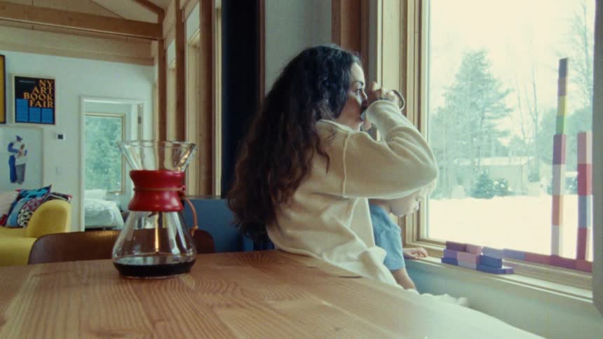 A woman with long dark curly hair holds a mug and stands near a large window looking out with a child beside her the child is playing with colorful blocks on the window ledge the interior is brightly lit with sunlight streaming through the window