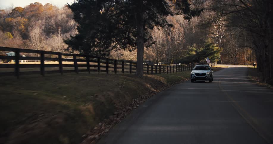 A gray car drives along a winding road with a wooden fence and a forest with bare trees visible on the side of the road