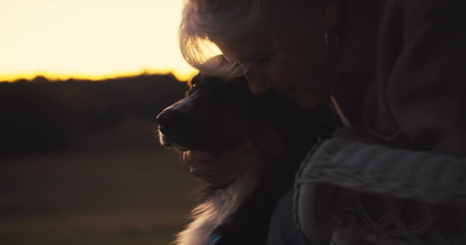An elderly woman sits on porch hugging and petting her dog as sun sets in the distance