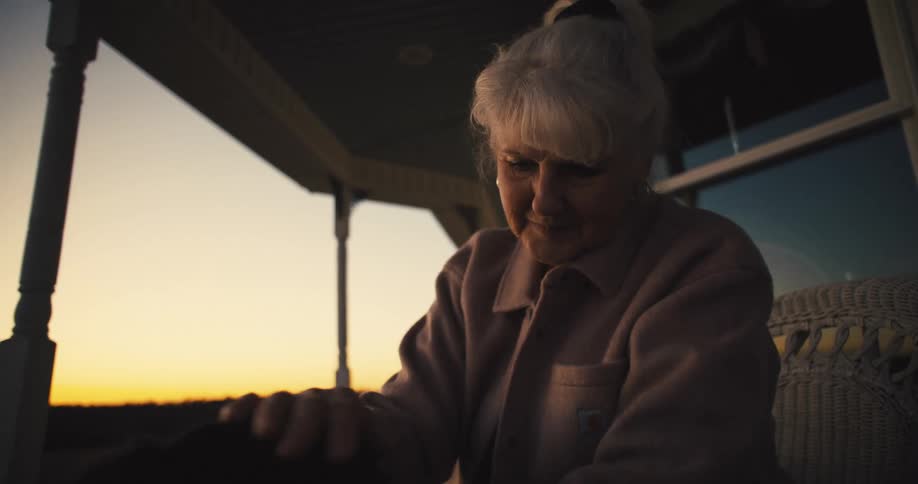 An elderly woman sitting in a chair in front of her house petting her dog