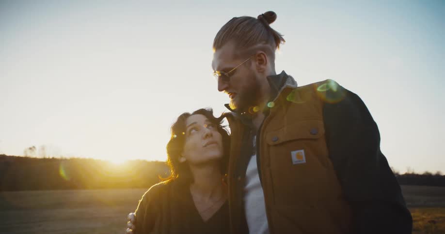 Based on the images from the video a man and woman are standing in a field at sunset the man who has a beard and sunglasses is wearing a vest and has his arm around the woman the woman is looking up at him while he looks into the distance with the s