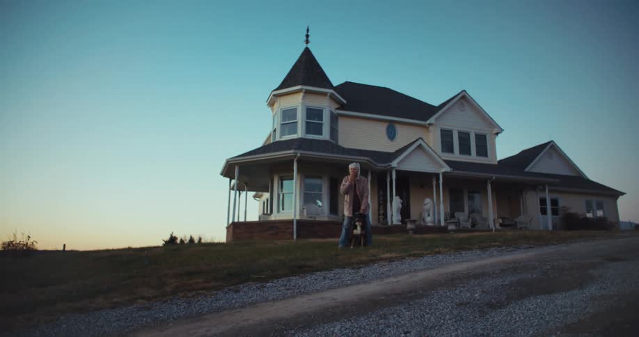 An elderly man in front of his house holding a dog saying goodbye to a car