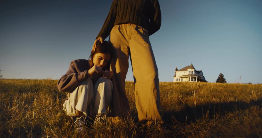 A tall person is standing over and petting the head of a girl who is crouched in a field of dry light brown grass with a white house on a hill visible in the background against a clear blue sky