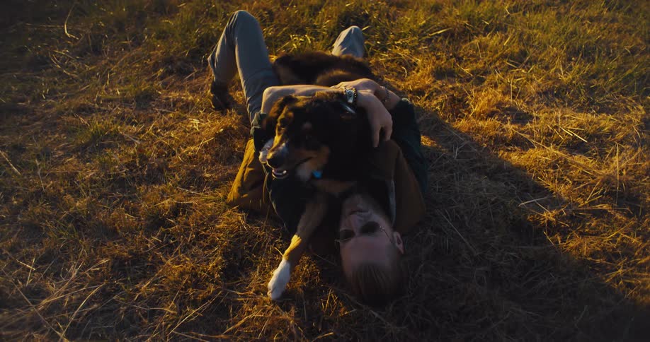 A young man pets and plays with his dog while lying on a field outdoors at sunset
