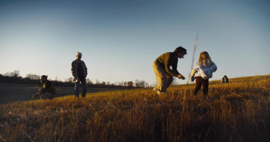 A man a woman and a child are playing with a black and white dog in a dry grassy field with a person standing nearby in the background under a blue sky