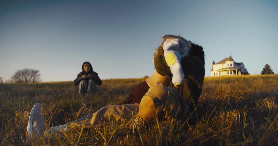 Based on the images from the video a woman with dark wavy hair is holding and comforting a young girl with blonde hair who is lying on the ground a second person is sitting in the background further up the hill with a large white house visible in th