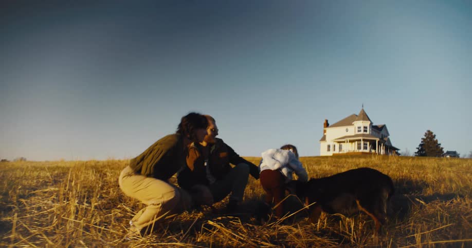 A family of threea young couple and their toddlerplays with their dog on a grassy field at midday with a building visible in the distance