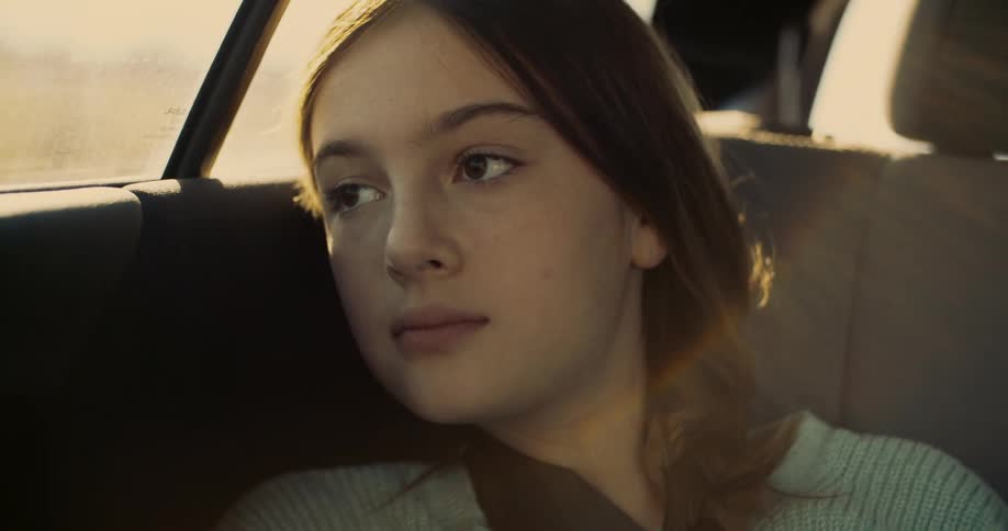 A teenage girl with long brown hair looks out the window of a car with the bright sunlight from outside casting a warm glow on her face