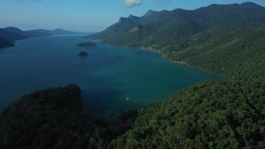 A vast body of deep blue water with a small sailboat is surrounded by lush green hills covered in trees with a coastal town visible along the shore in the distance