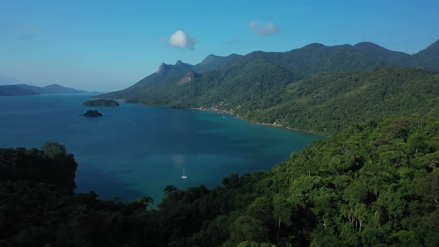 The video shows an aerial view of a stunning natural landscape featuring a large body of clear blue water surrounded by lush green mountains a small white boat is visible sailing gently on the tranquil water