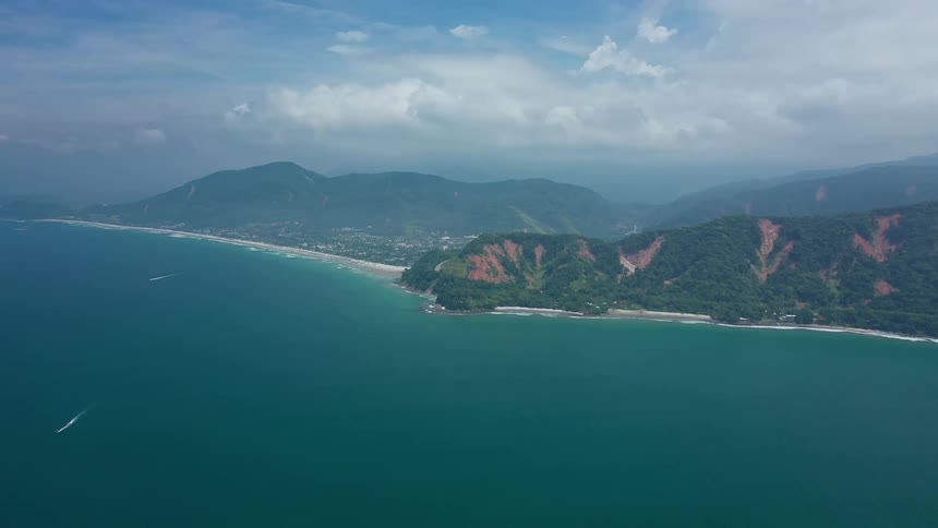 A high aerial view shows a long coastline with a town next to a green mountain range with visible landslides as a large body of deep blue water with small wakes from a couple of boats stretches out to the foreground under a partly cloudy sky