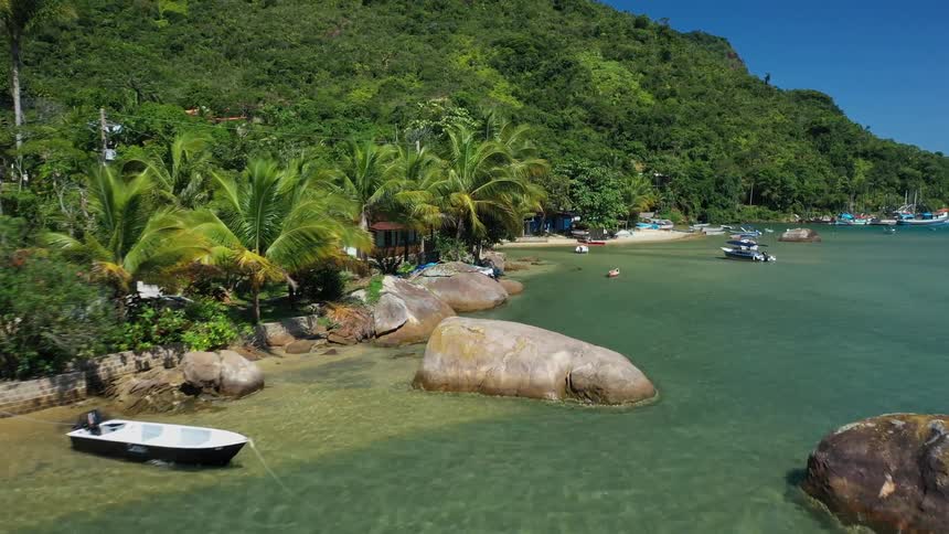 An aerial view of a small coastal town shows a green hillside covered in trees and residential buildings that lead down to a sandy beach with large rocks with a variety of boats and kayaks floating on a turquoise ocean