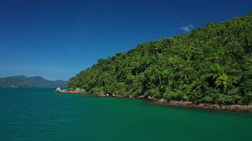 A bay of deep blue water is surrounded by a tropical island covered in green foliage and rocky outcrops on the right and a larger distant mountain on the left
