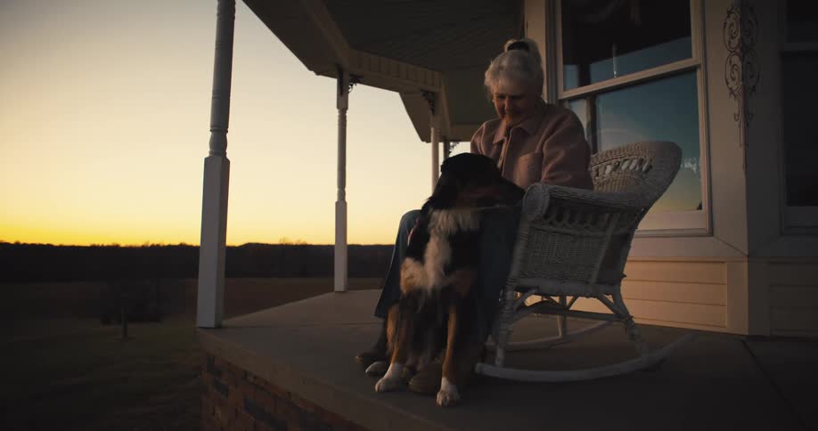 Older woman sits in a rocking chair on the porch of her rural home after dusk and pets her dog