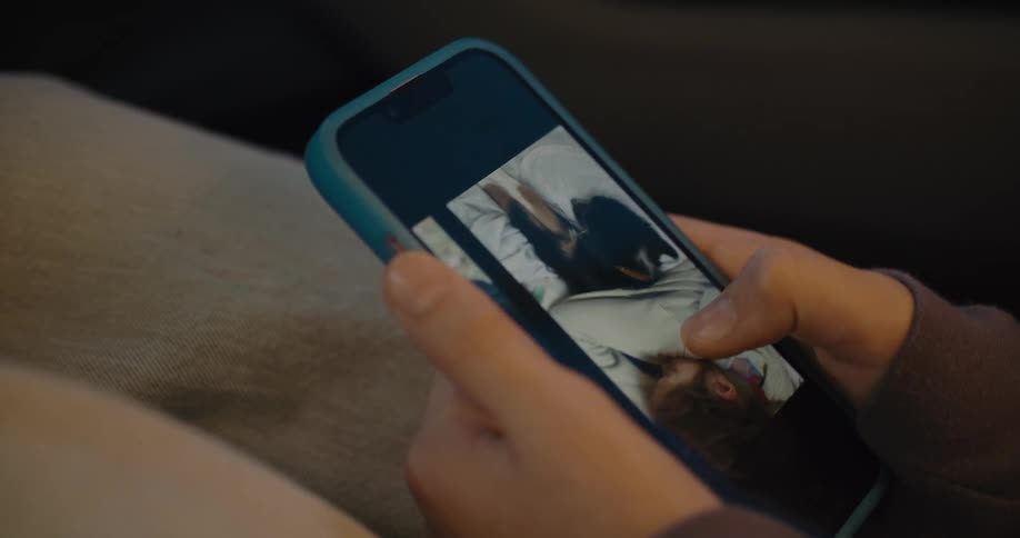 A close angled view shows a person holding a blue cellphone with their thumb scrolling through various photos of a black and white dog
