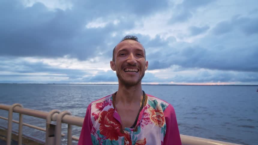 A man with a wide smile and a partial goatee stands on a waterfront pier wearing a pink shirt with a colorful floral pattern with a calm sea and a cloudy blue and orange sky visible behind him