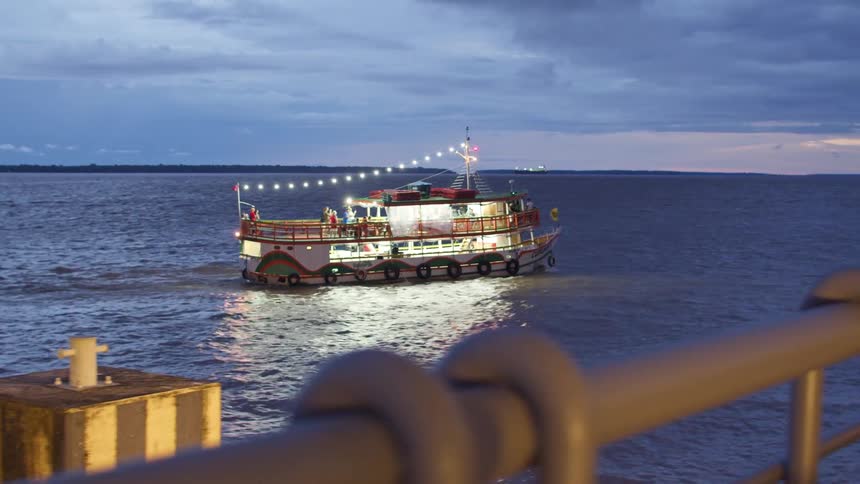 Based on the images from the video a large passenger ferry decorated with lights is sailing on a wide river at dusk the sky is a cloudy grey and the water reflects the lights from the boat and the dim light from the sky