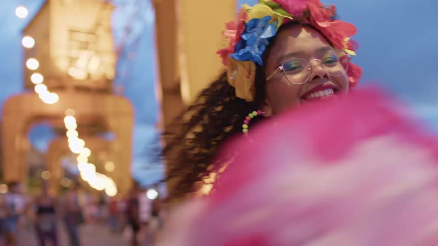 A woman with dark curly hair and glasses wearing a white lace top a colorful floral headband and bright colorful necklaces and earrings is smiling as she dances on a pier at sunset