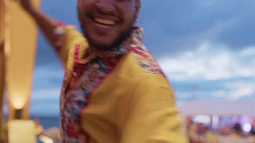 Based on the images from the video a man with a beard and a yellow floral shirt is smiling widely he is standing outdoors possibly at a market with a blue sky and white clouds in the background the man raises one arm and moves it back and forth as if