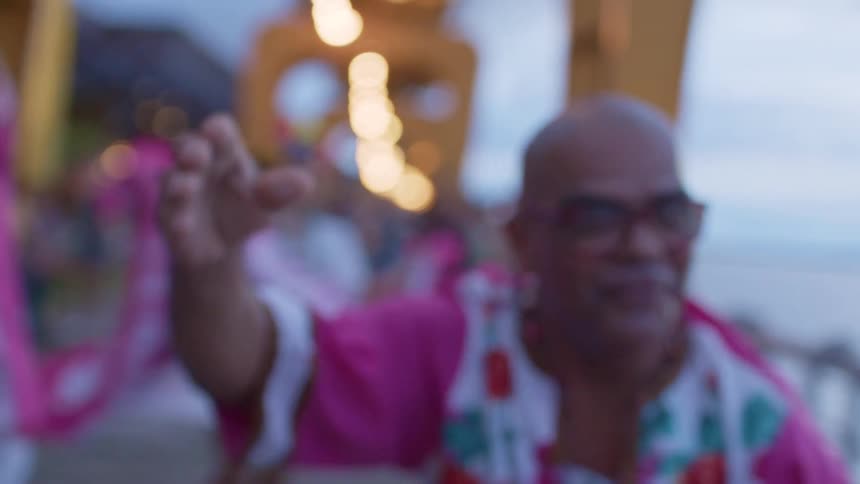 A closeup shot focuses on a balding man with redrimmed glasses a necklace and a pink floral shirt he is smiling and dancing with other dancers and a city background visible behind him