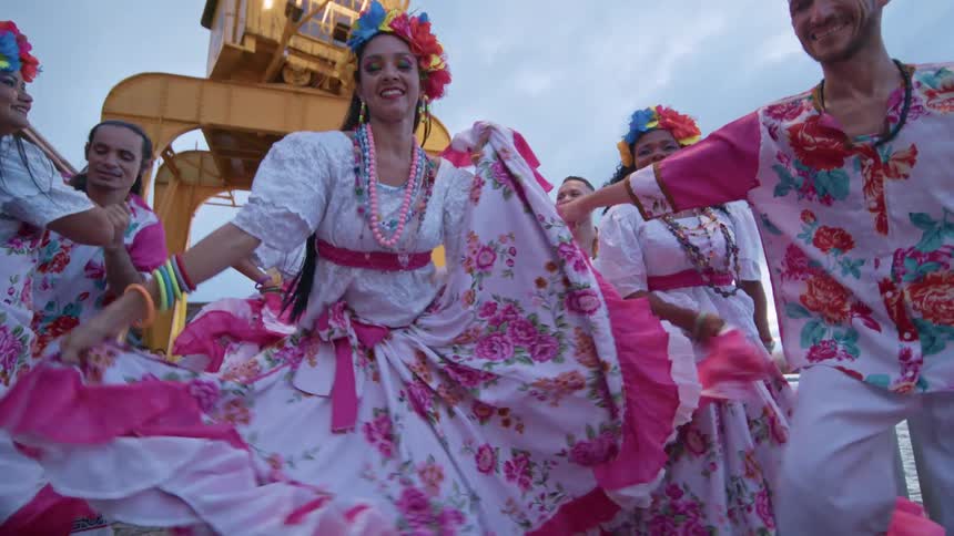 A group of dancers dressed in colorful traditional clothing are performing a lively dance outdoors the women are wearing white dresses with pink floral patterns and bright pink ruffled underskirts
