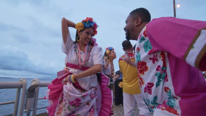 A group of people dressed in matching cultural attire dances in celebration while others play musical instruments at the dock at midday