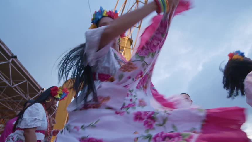 In a lively outdoor setting a group of people is seen dancing dressed in colorful traditionalstyle clothing the women wear long flowing skirts with floral patterns and bright pink underskirts while the men wear matching floral shirts and white pant