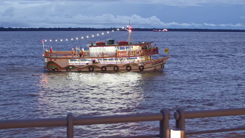 Seen from a waterfront at dusk a twostory party boat named curumim decorated with string lights floats on a wide river with people on its top deck