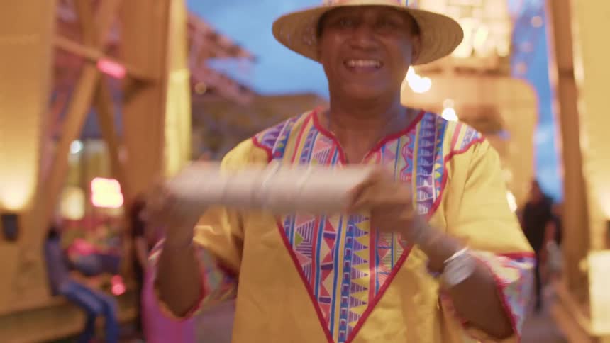 A man wearing a yellow and patterned shirt and a woven hat with a colorful band is smiling and shaking a light colora man wearing a yellow and patterned shirt and a woven hat with a colorful band is smiling and shaking a light colored cylindrical instrume