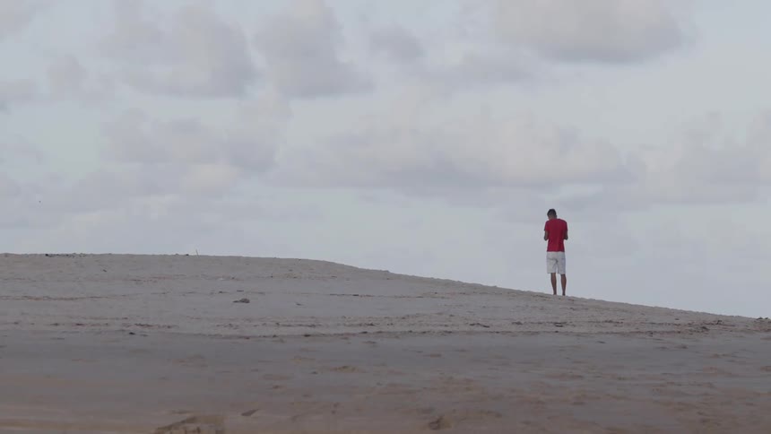 A person in a red shirt and white shorts stands on a sandy hill and then begins to walk away