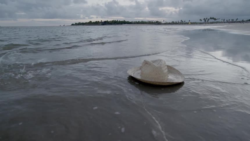 A straw hat lying in the wet sand at the waters edge on a beach and then a person walks up and bends down to pick it up