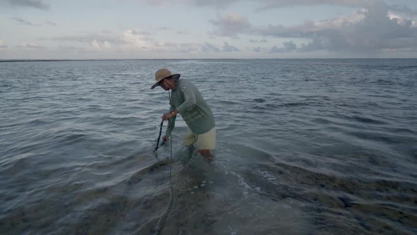 A man wearing a sun hat stands in the ocean near the beach setting a fishing line at midday