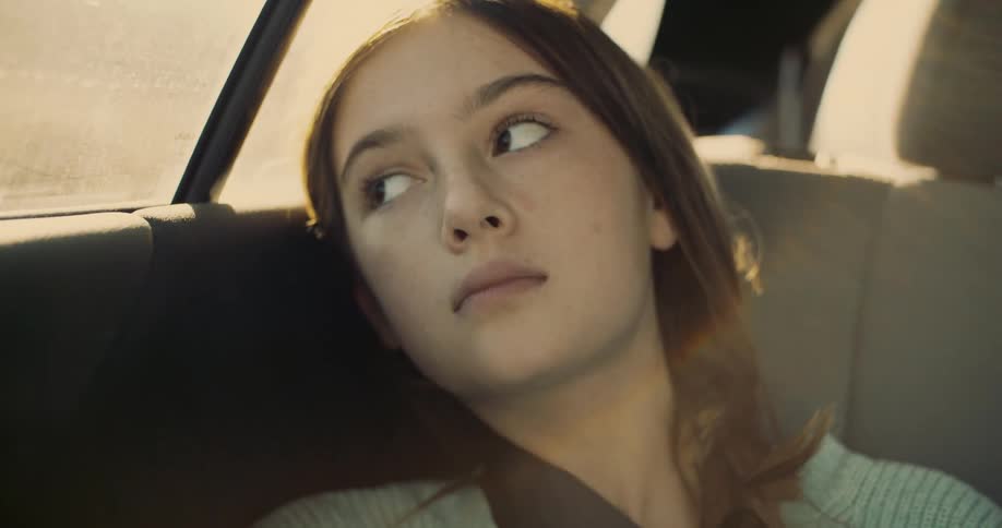A girl sitting in the back of a car looking out the window with a pensive and distant expression