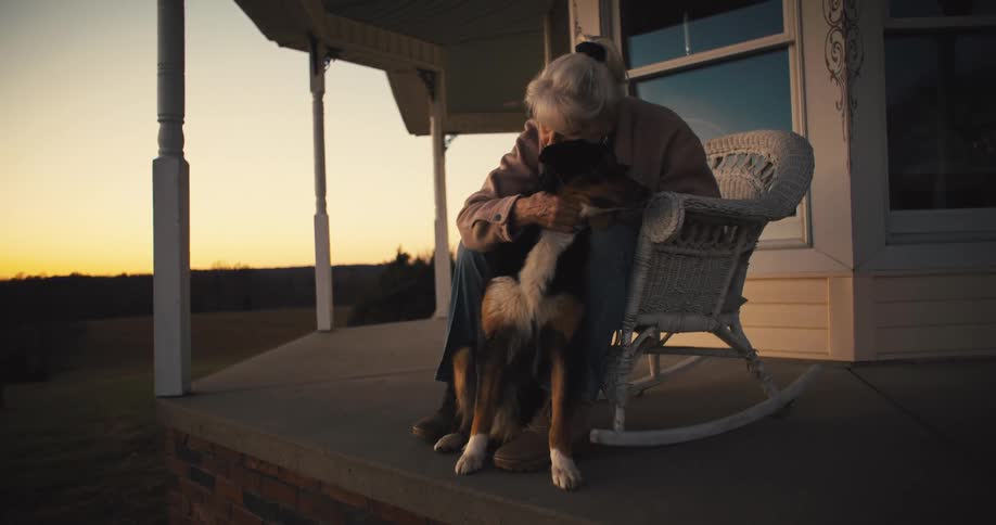 A woman with white hair in a bun is sitting on a front porch in a rocking chair lovingly petting a black and tan dog as the sun sets behind them