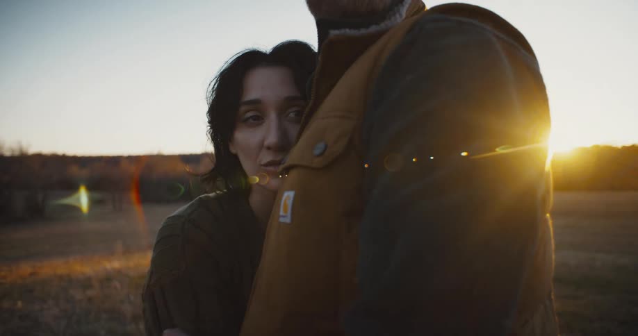 A woman with dark hair looks up at a man in a brown vest then embraces him as the sun sets behind them in a field