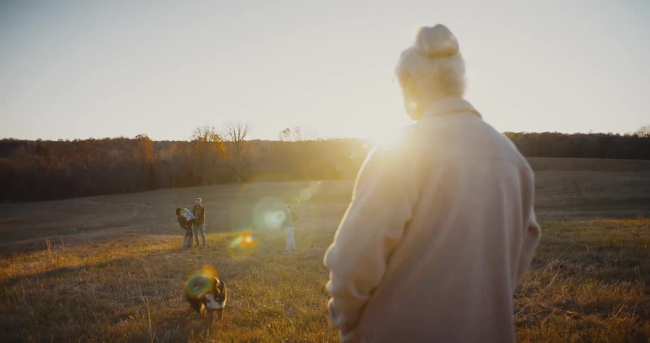 Woman watches family members and a dog in an open field at sunset