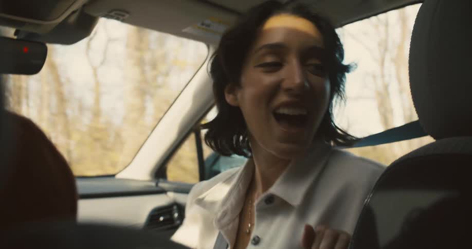 A woman with short dark hair is in the passenger seat of a car smiling and looking back towards the back seat with his head turned
