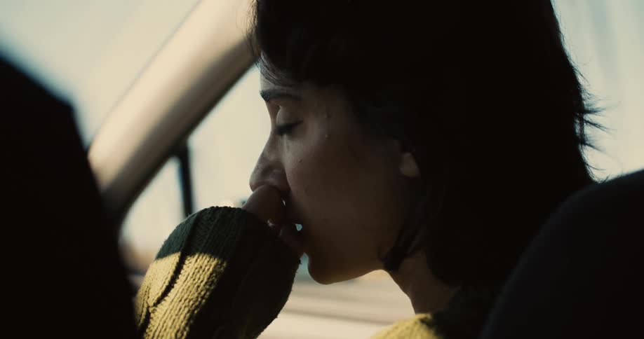 A young woman sits in the passenger seat of a car talking to the driver as the car moves at midday