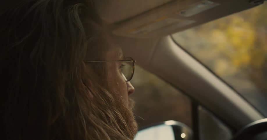 A man with long hair wearing glasses drives a car at midday with trees lining the roadside