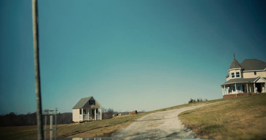 A vehicle is traveling up a winding dirt driveway toward a large light colored house with a turret and a porch while a smaller house and a leafless tree stand on the hillside to the left