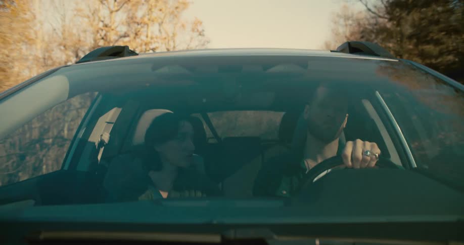 A young woman sits in the passenger seat of a car at midday having a conversation with the man driving
