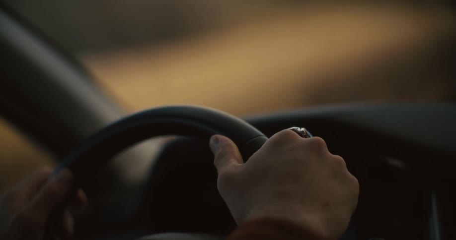 A person with light skin wearing a brown corduroy jacket and a ring on their right hand is holding the steering wheel of a car with both hands and driving forward
