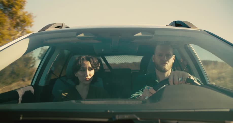 A wide shot through a cars windshield shows a man and a woman sitting in the front seats the man is driving and speaking while the woman looks at him intently listening