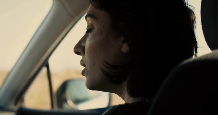 A woman with short dark hair is shown in a side view looking out the passenger window of a moving car with a saddened expression