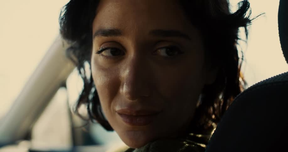 A young woman sits in the passenger seat of a moving car looking at the back seat as she talks to someone with sadness in her eyes then wipes away her tears at midday
