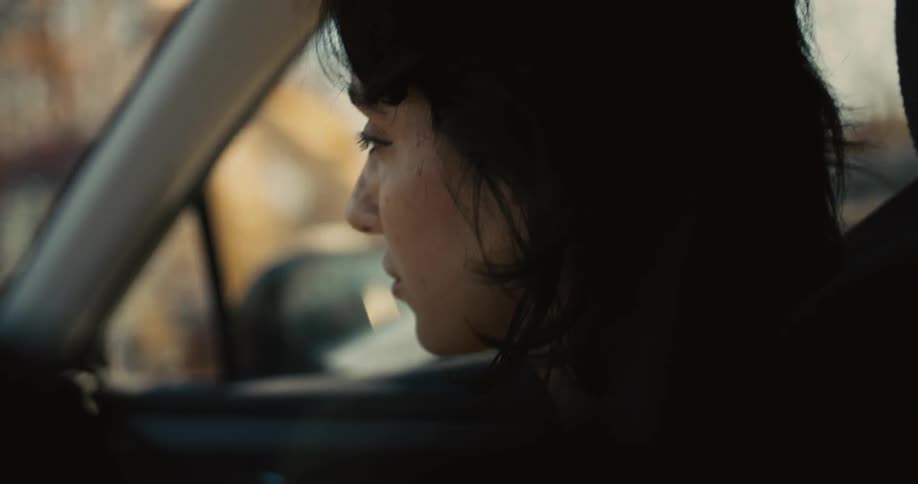Based on the video a closeup shot shows a persons face from the side while they are driving a car the person is looking out the passenger window and their face is lit by the warm glow of the sun the window shows trees and a building passing by